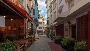 Una strada acciottolata fiancheggiata da edifici, alcuni con persiane rosse e altri con insegne. Sono presenti aree con posti a sedere all'aperto, tavoli e sedie, e vasi con piante lungo il percorso.