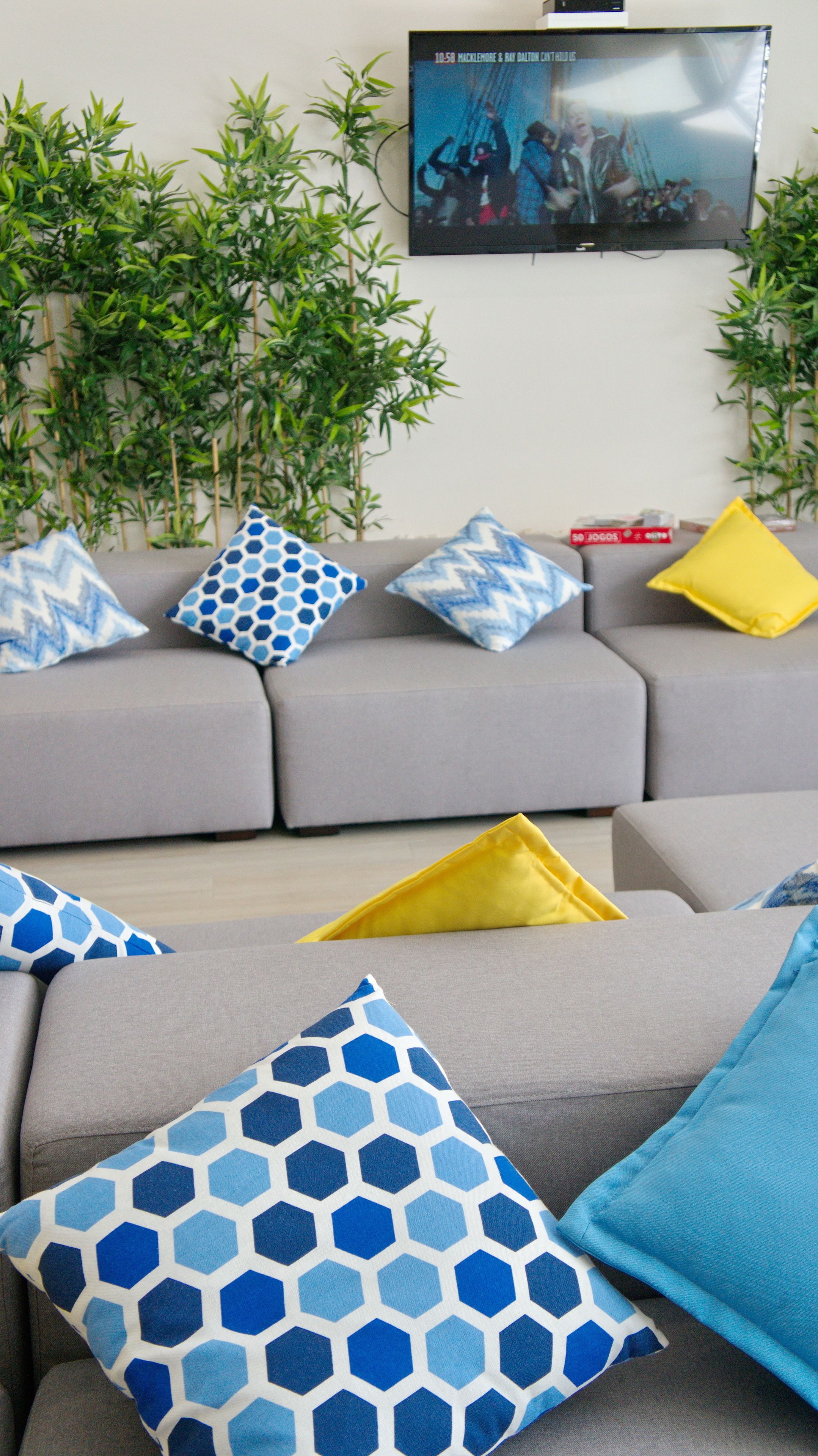 A living room with a grey sofa, blue and yellow patterned cushions, and a flat-screen TV mounted on the wall.