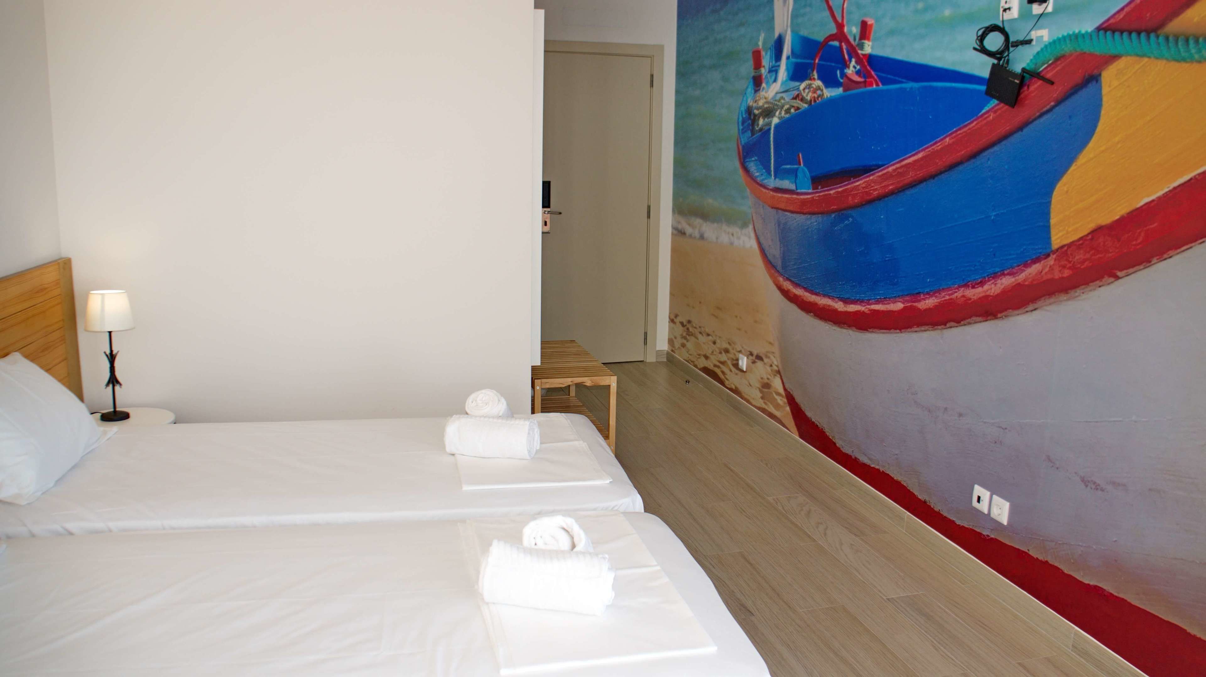 A hotel room with two beds, a wooden headboard, a bedside table with a lamp, and a mural of a boat on the wall.