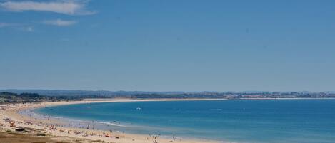 A wide beach with clear blue water, sandy shore, and distant cityscape.
