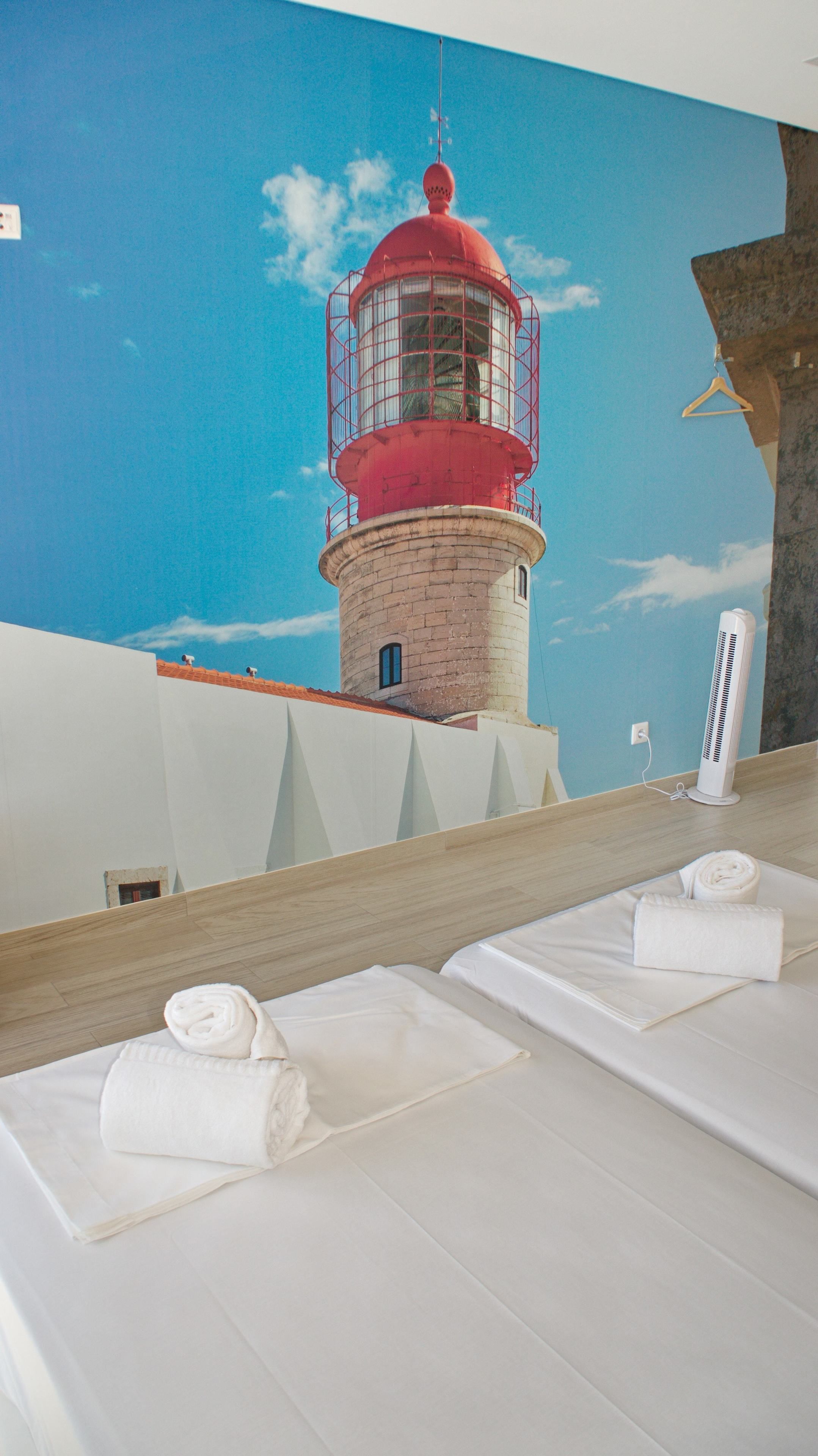 A room with a mural of a lighthouse, a bed with a towel, and a white fan.