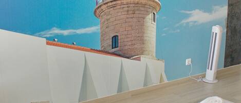 Habitación con un mural de un faro, una cama con una toalla y un ventilador blanco.