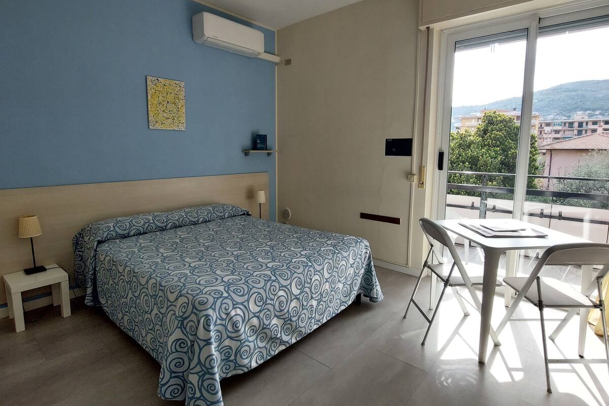 Ein Schlafzimmer mit einem Bett, einem kleinen Nachttisch, einem Sessel und einem Fenster mit Blick auf Gebäude und Bäume.