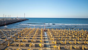 A beach with rows of sun loungers and umbrellas, a pier with boats, and a lighthouse in the background. - Residence La Palma (Andora)
