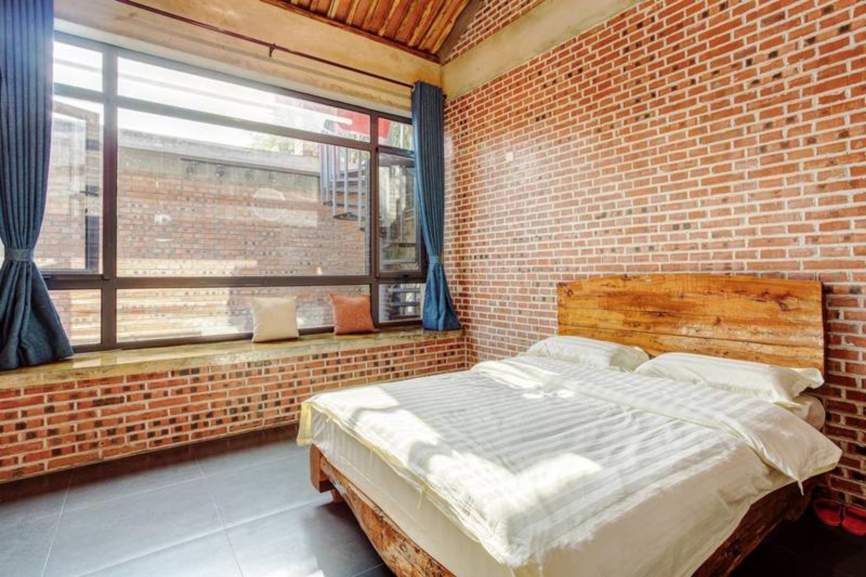 A room with a brick wall, a large window with curtains, a wooden headboard, and a bed with white linens.