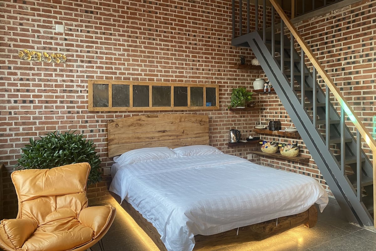 Ein Schlafzimmer mit Backsteinwand, Holz-Kopfteil, einem Bett mit weißer Bettwäsche, einem Ledersessel und einer Treppe mit Metallgeländer.