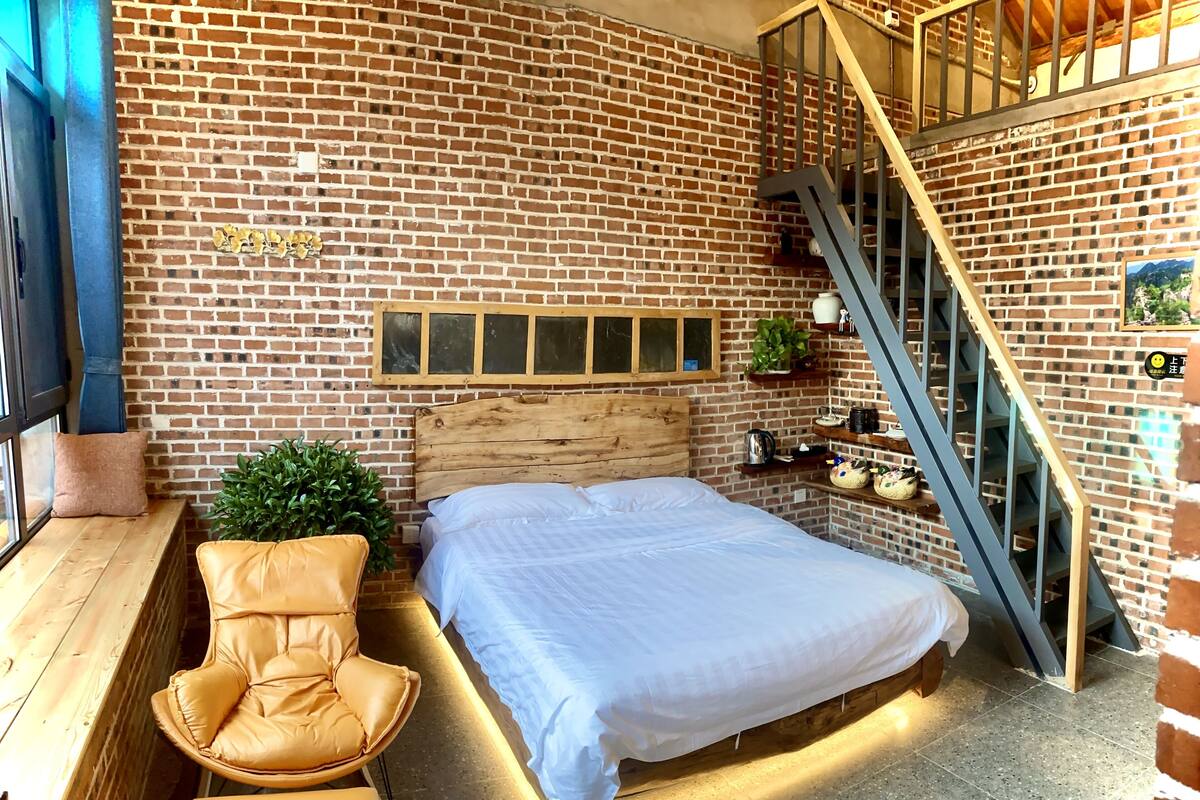 Ein Schlafzimmer mit Backsteinwand, einem hölzernen Bett, einer Bank, einem Stuhl und einer Treppe.