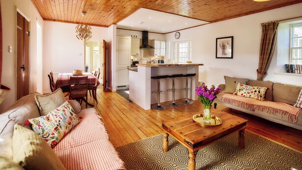 A cozy living room with a wooden ceiling, a sofa, a coffee table, and a dining area with a table and chairs. - Mauldslie Hill Cottage (Gorebridge)
