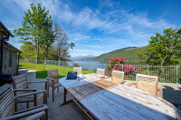 A wooden deck with chairs and a table, overlooking a lake and hills.