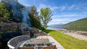 An outdoor seating area with a stone wall, a wooden table, and chairs, overlooking a lake and hills. - Taymouth Marina - Port Bane (Aberfeldy)