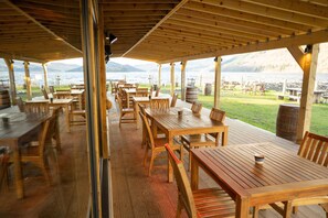 An outdoor wooden deck with tables and chairs, overlooking a lake with mountains in the background.