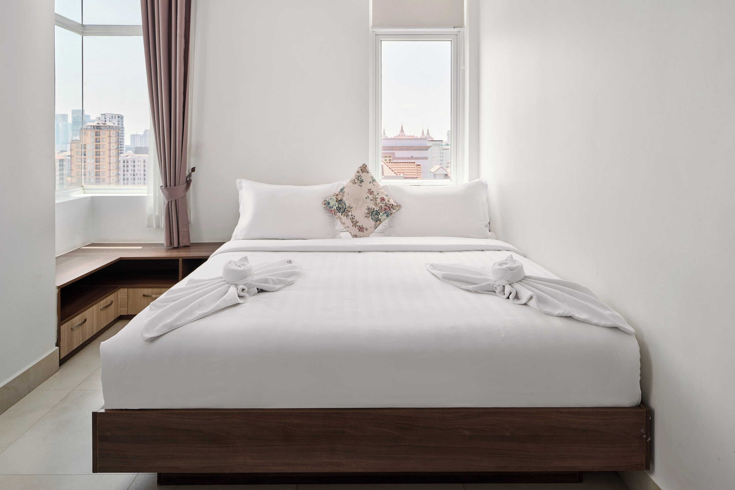 A modern hotel room with a large bed, a view of the city through a window, and a wooden nightstand with a lamp.