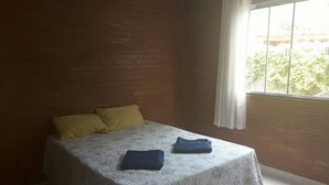 A single bed with a floral bedspread and two pillows, a window with curtains, and a view of greenery outside. - Casa Temporada Campeche (Florianópolis)