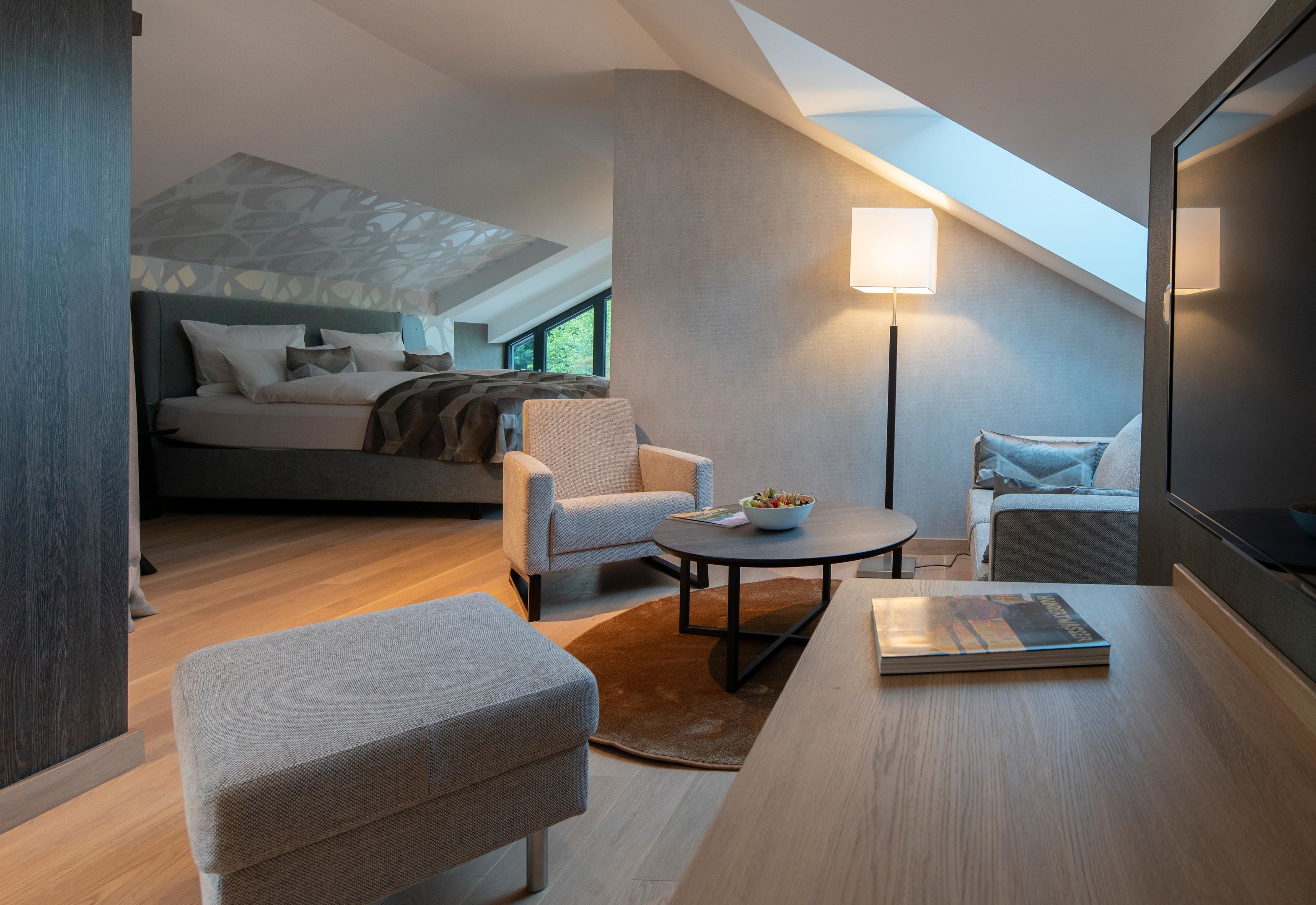 Ein modernes Hotelzimmer mit Schlafpodest, einer Sitzecke inklusive rundem Tisch und einem großen Fenster, das natürliches Licht hereinlässt.