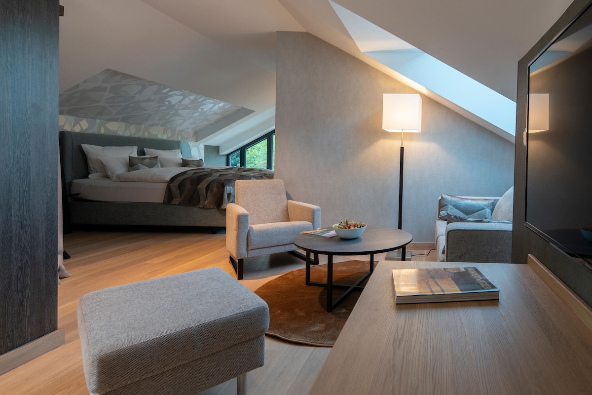 A modern hotel room with a loft bed, a seating area with a round table, and a large window providing natural light.