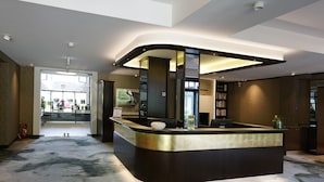 A modern reception desk with a curved design, a glass partition, and a digital screen. There is a fire extinguisher on the left and a bookshelf with books to the right. - Essensio Hotel Düsseldorf (Erkrath)
