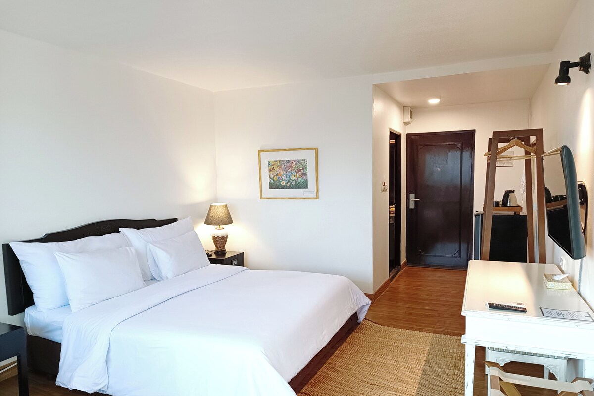 A hotel room with a bed, bedside lamp, a desk with a mirror, and a door leading to another room.