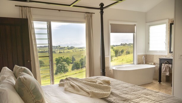 A bedroom with a large bed, a view of a green landscape, a bathtub, and a window. - The best Byron Bay has to offer - stunning views, total luxury and style.   (Coopers Shoot)