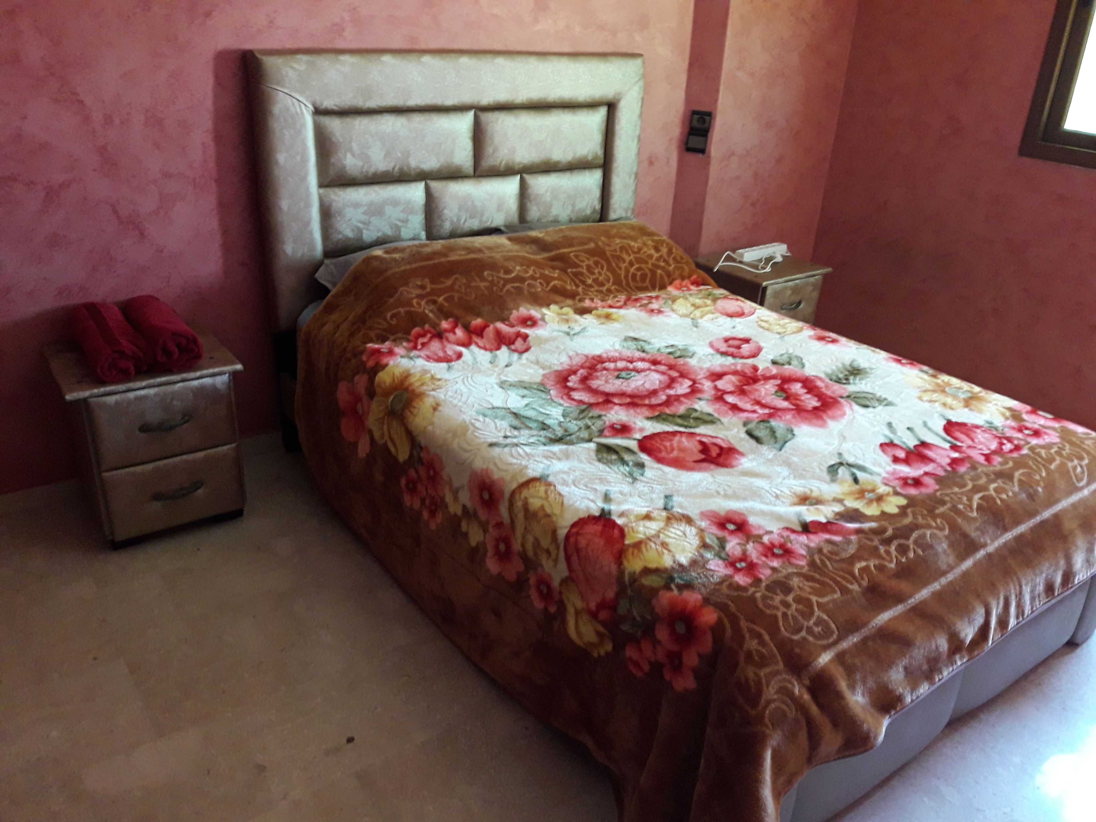 A bedroom with a floral bedspread, a headboard, a nightstand with a phone, and a red towel on the side table.