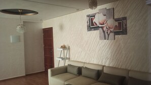 Un salon avec un canapé beige, une porte en bois, un luminaire fixé au mur et une œuvre d’art florale encadrée.