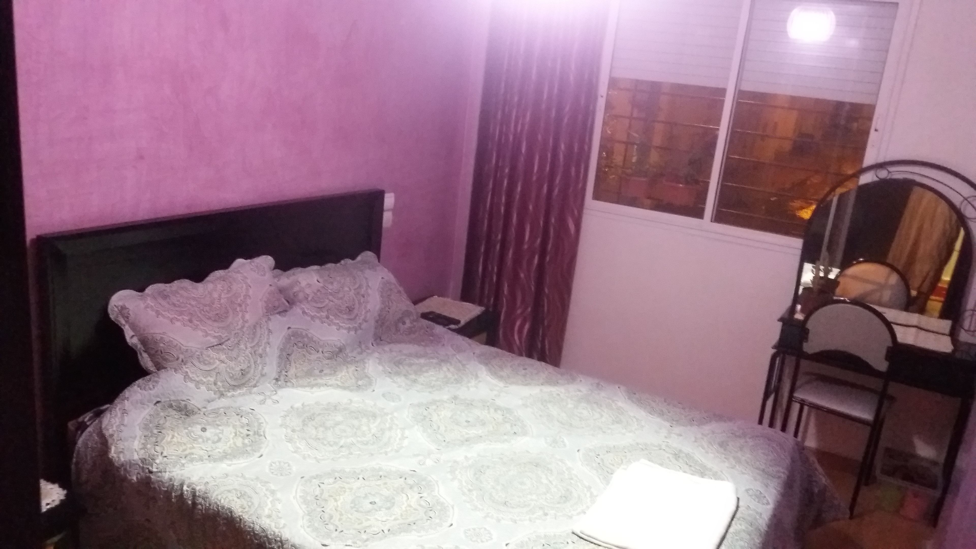 Un dormitorio con cama, mesita de noche, silla y ventana con cortinas.