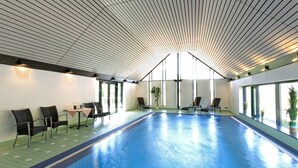 An indoor swimming pool with a modern ceiling design, seating area with tables, and large windows providing natural light. - Hotel Hirschen Glottertal (Glottertal)