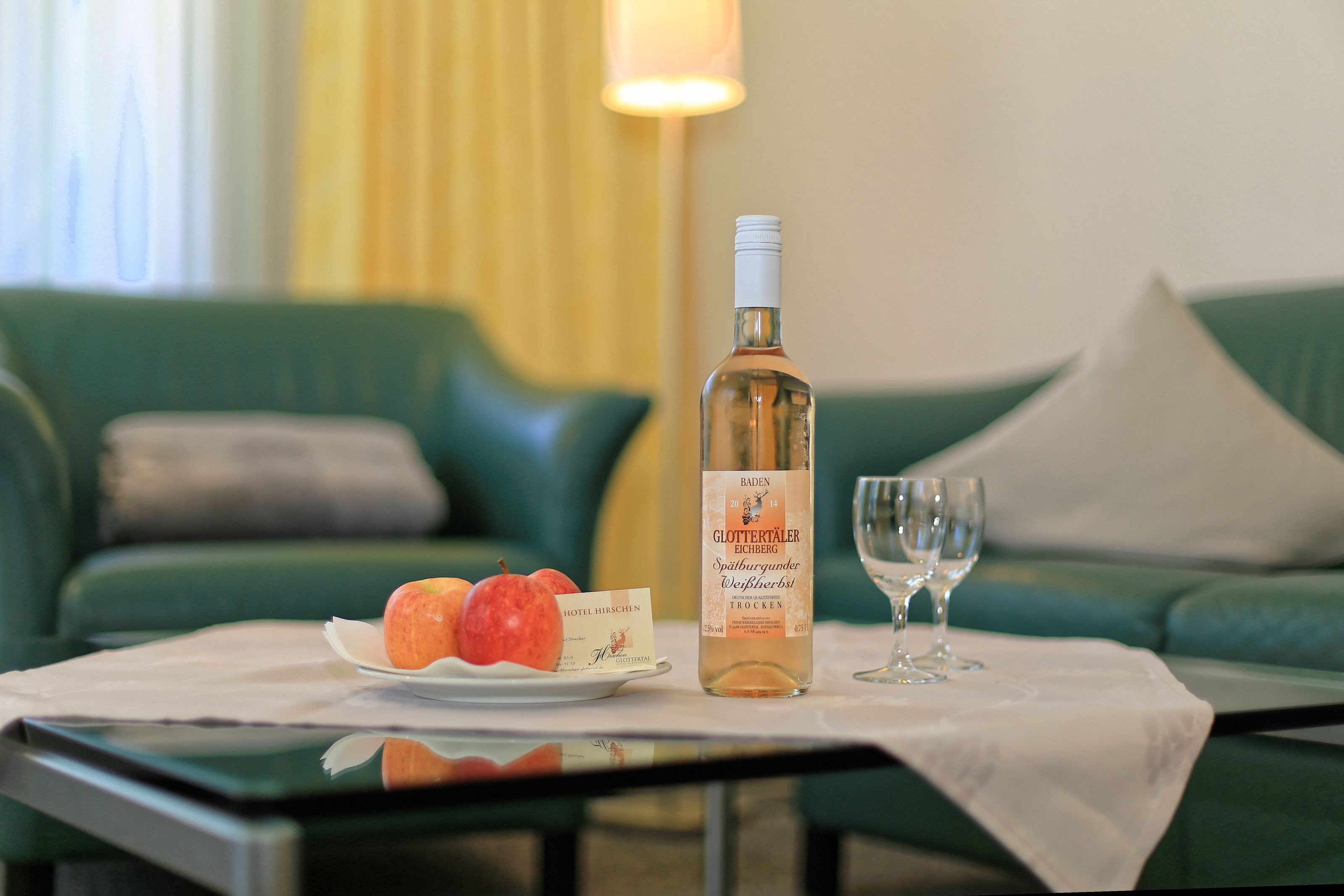 A table with a bottle of wine, a glass, and a plate of apples in a hotel room.