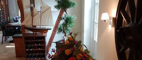 Una mesa de madera con un jarrón de flores, un modelo de barco y una botellera en una habitación con vigas a la vista y una rueda grande.