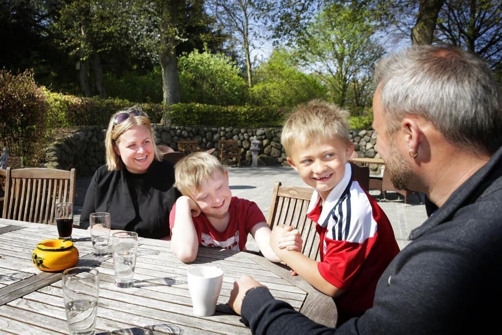 Eine Familie speist an einem Tisch im Freien, mit einer Steinwand und Grünpflanzen im Hintergrund.