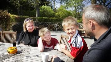 Eine Familie speist an einem Tisch im Freien, mit einer Steinwand und Grünpflanzen im Hintergrund.