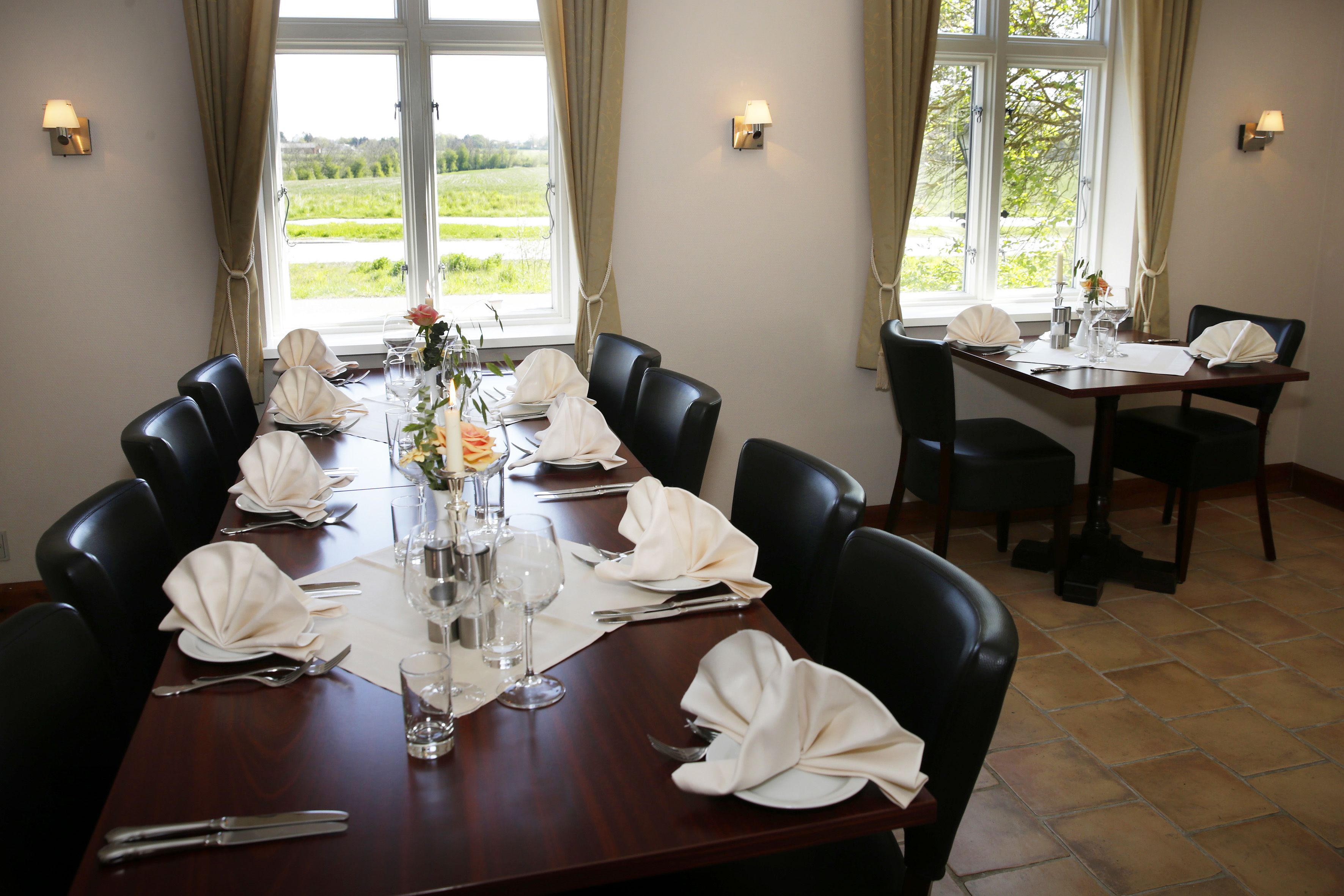 A dining room with a long table set for a meal, surrounded by chairs, with a view of greenery outside.