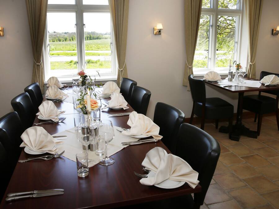 A dining room with a long table set for a meal, surrounded by chairs, with a view of greenery outside.