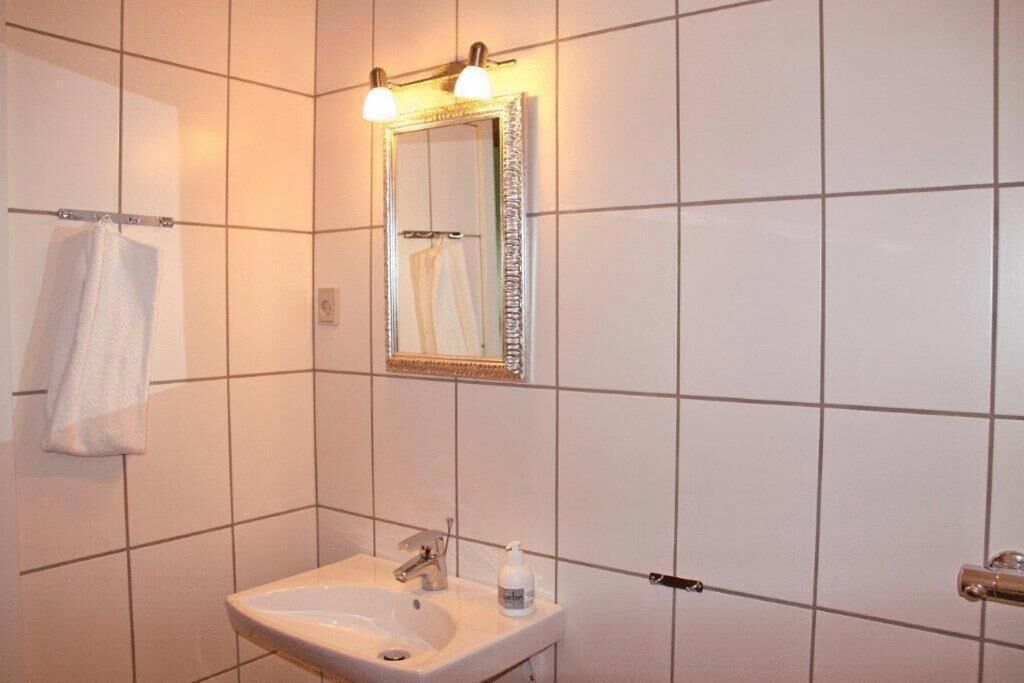Un baño con paredes de azulejos blancos, un lavamanos con espejo y una toalla colgada en un perchero.