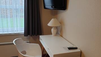 A hotel room with a white desk, a television mounted on the wall, a white chair, a brown leather chair, and a window with curtains.