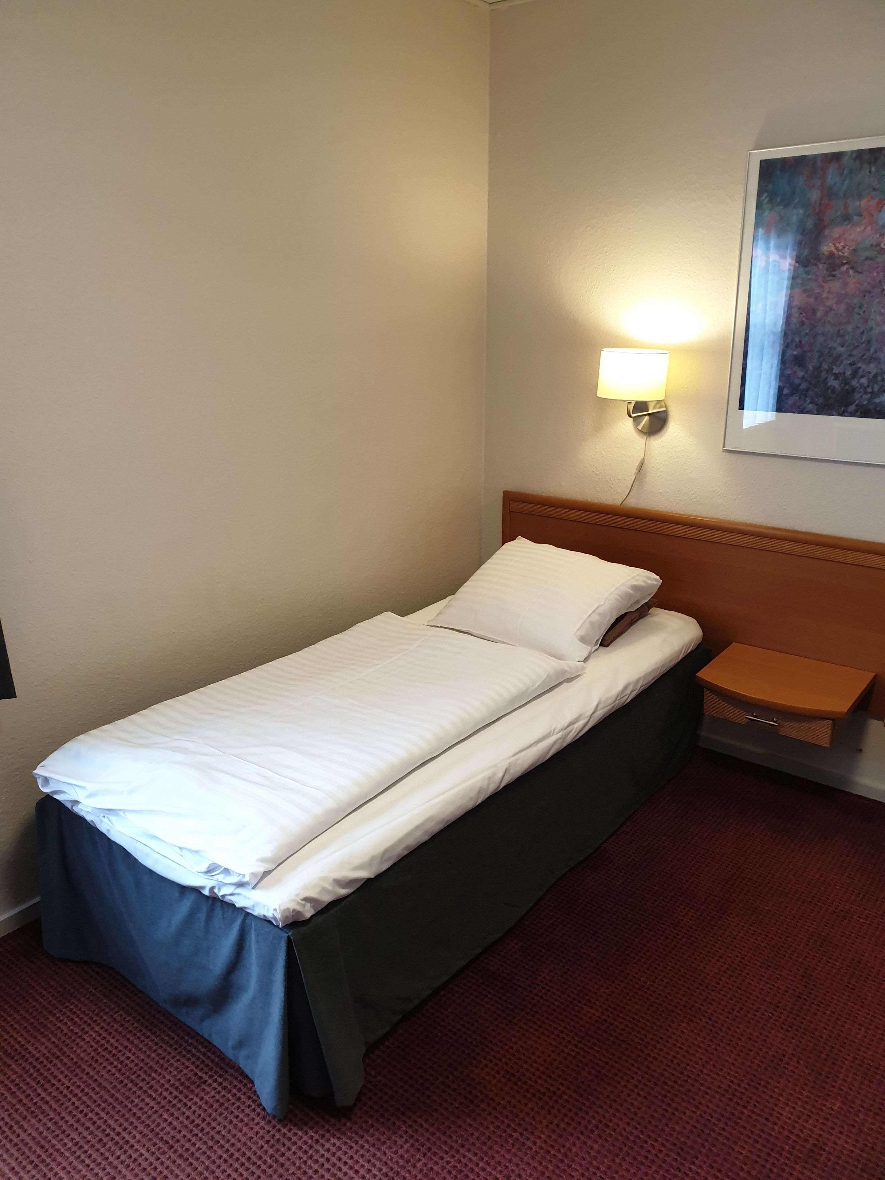 Un letto singolo con una testiera in legno, un comodino con una lampada e un quadro appeso alla parete.