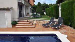 Une piscine avec des marches menant à une maison, deux chaises longues et un jardin bien entretenu.