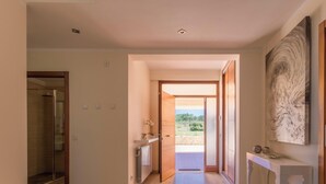 Corredor com piso de madeira, box de vidro para chuveiro e vista para o exterior com áreas verdes.