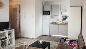 Un salon moderne avec un téléviseur, une table basse et un canapé. On aperçoit une cuisine à l’arrière-plan, avec des meubles blancs, un four à micro-ondes et un réfrigérateur.