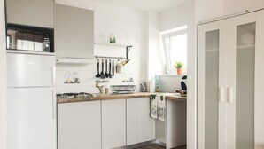 Une cuisine moderne avec des meubles blancs, un four encastré et une cuisinière à gaz. On y trouve des ustensiles de cuisine suspendus et une fenêtre avec une plante posée sur le rebord.
