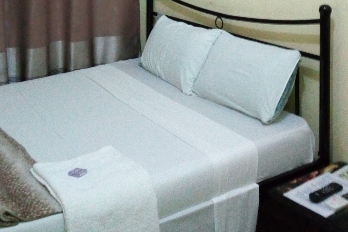 A bed with a white mattress and pillows, a dark metal headboard, a brown curtain with a pattern, a ceiling fan, and a nightstand with a remote control.
