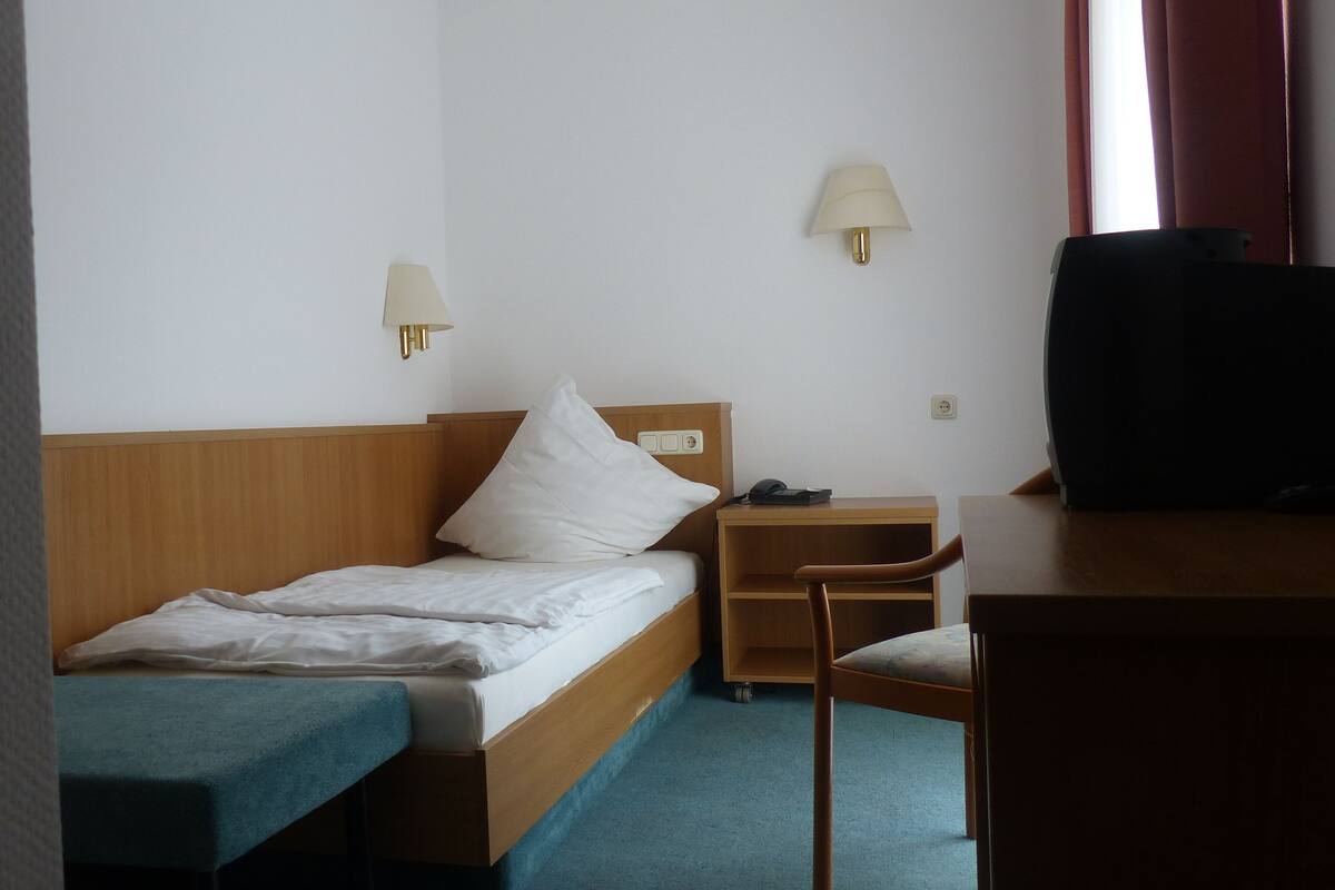 A hotel room with a single bed, a desk with a chair, a television, and two wall-mounted lamps.