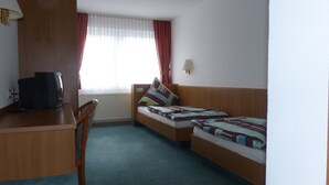 Ein Hotelzimmer mit Holzschreibtisch, Fernseher, zwei Betten mit farbenfroher Bettwäsche, einem Fenster mit leichtem Vorhang und einer Wandlampe.