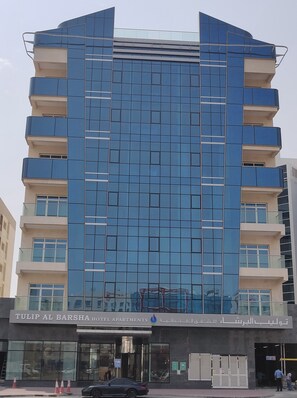 A modern hotel building with a glass facade, multiple balconies, and a sign indicating it is Tulip Al Barsha Hotel Apartments.