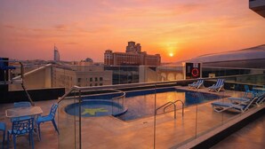 A rooftop pool with lounge chairs, a glass balustrade, and a sunset view of city buildings.