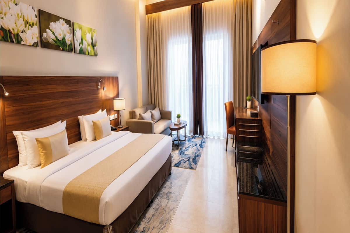 A hotel room with a large bed, a desk, a chair, a TV, and a lamp.