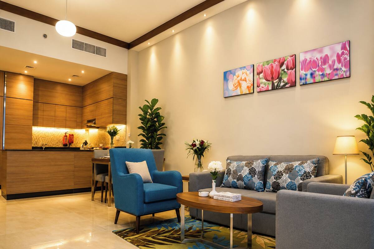 A modern living room with a blue armchair, grey sofa, and a coffee table. There are three wall-mounted flower paintings, a kitchen area with wooden cabinets, and a dining table with chairs.