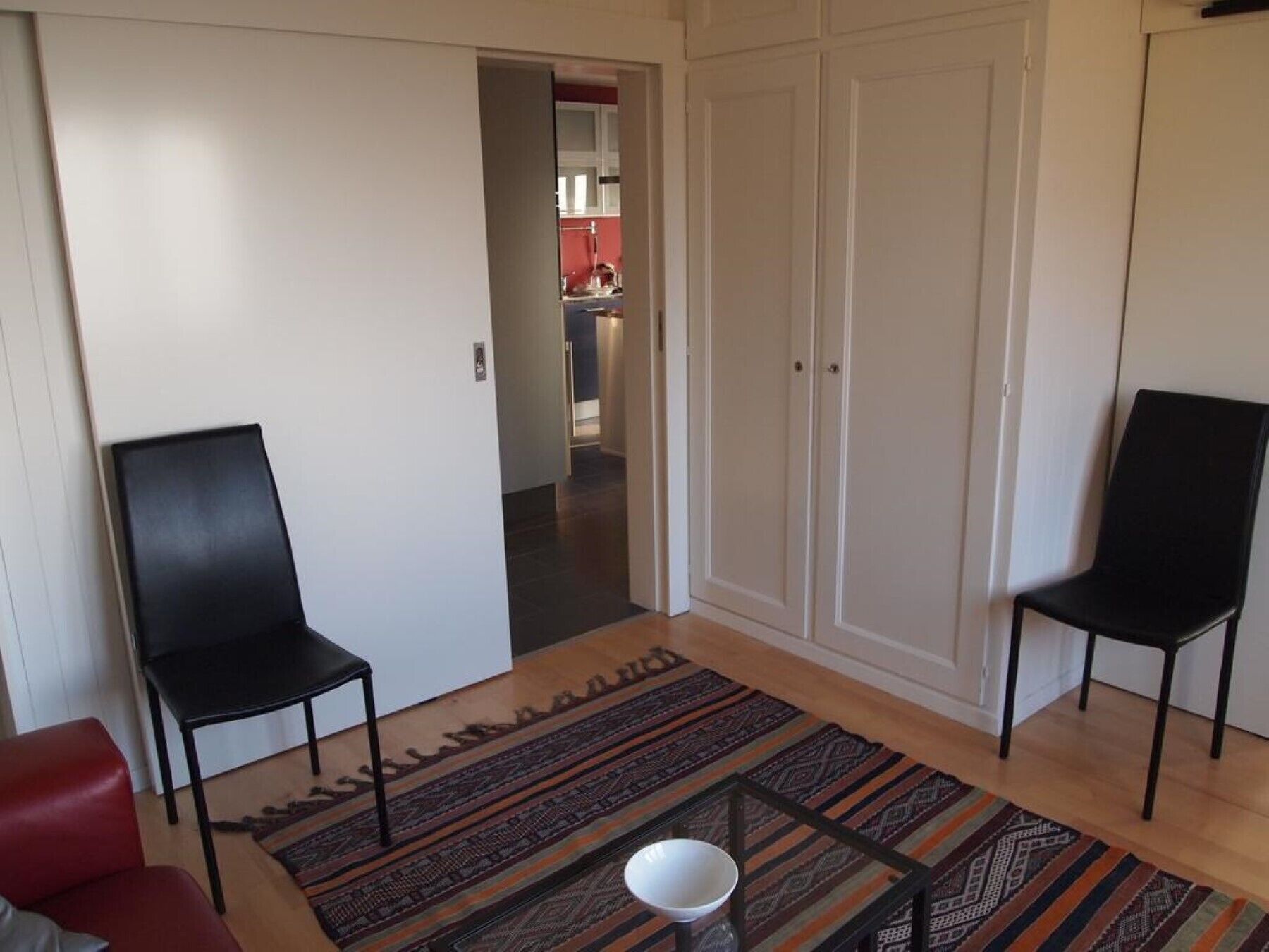 Una sala de estar con dos sillas negras, un sofá rojo, un tapete estampado y una mesa de vidrio.