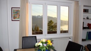 Zona pranzo con tavolo, sedie, un vaso di fiori, una finestra vista montagna e un termosifone.