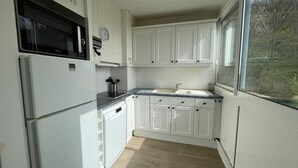 Una cocina con armarios blancos, microondas, lavamanos y ventana con vista a árboles.