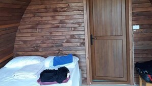 A small, cozy room with a sloped ceiling and wooden walls. It features a single bed with a white mattress and a blue blanket, a wooden door, and a small table with a chair. - Metis Bungalow (Ayvacik)
